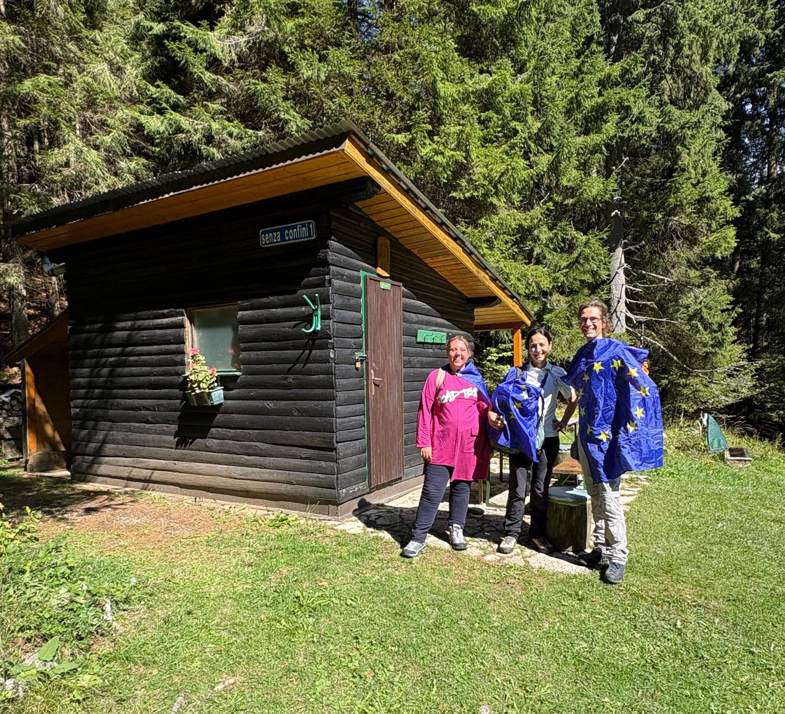 Il team di Europe Direct Trieste in posa con le bandiere dell'Unione Europea davanti a una struttura in legno immersa nel bosco del Monte Forno, recante l'insegna "senza confini 1".