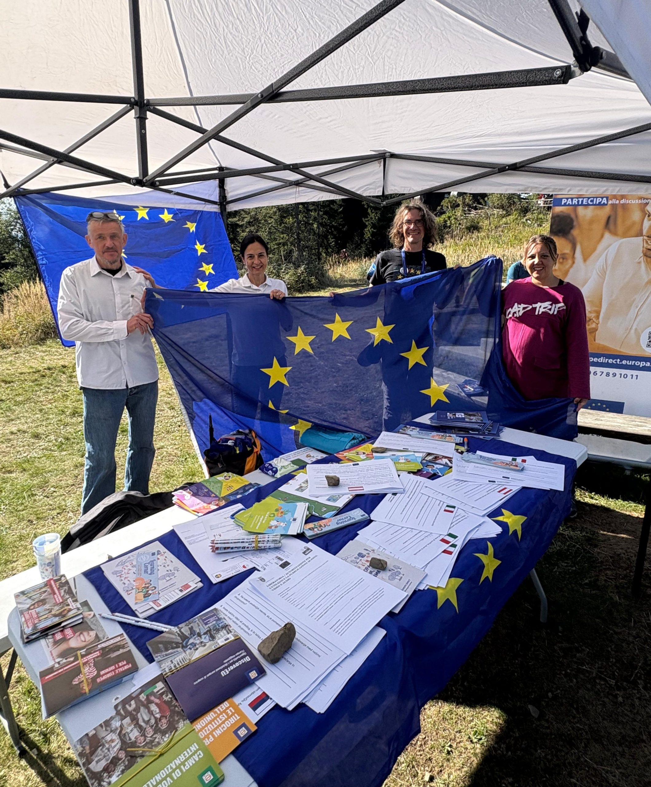 Il team di Europe Direct Trieste insieme al collega di Europe Direct Montagna Friulana sorridono sotto il gazebo informativo sul Monte Forno, sorreggendo la bandiera dell'Unione Europea sopra un tavolo allestito con brochure e materiali sulla mobilità giovanile.