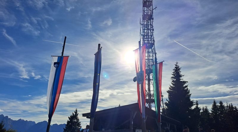 Bandiere di Slovenia, Unione Europea, Austria e Italia che sventolano sulla cima del Monte Forno sotto un cielo azzurro soleggiato.