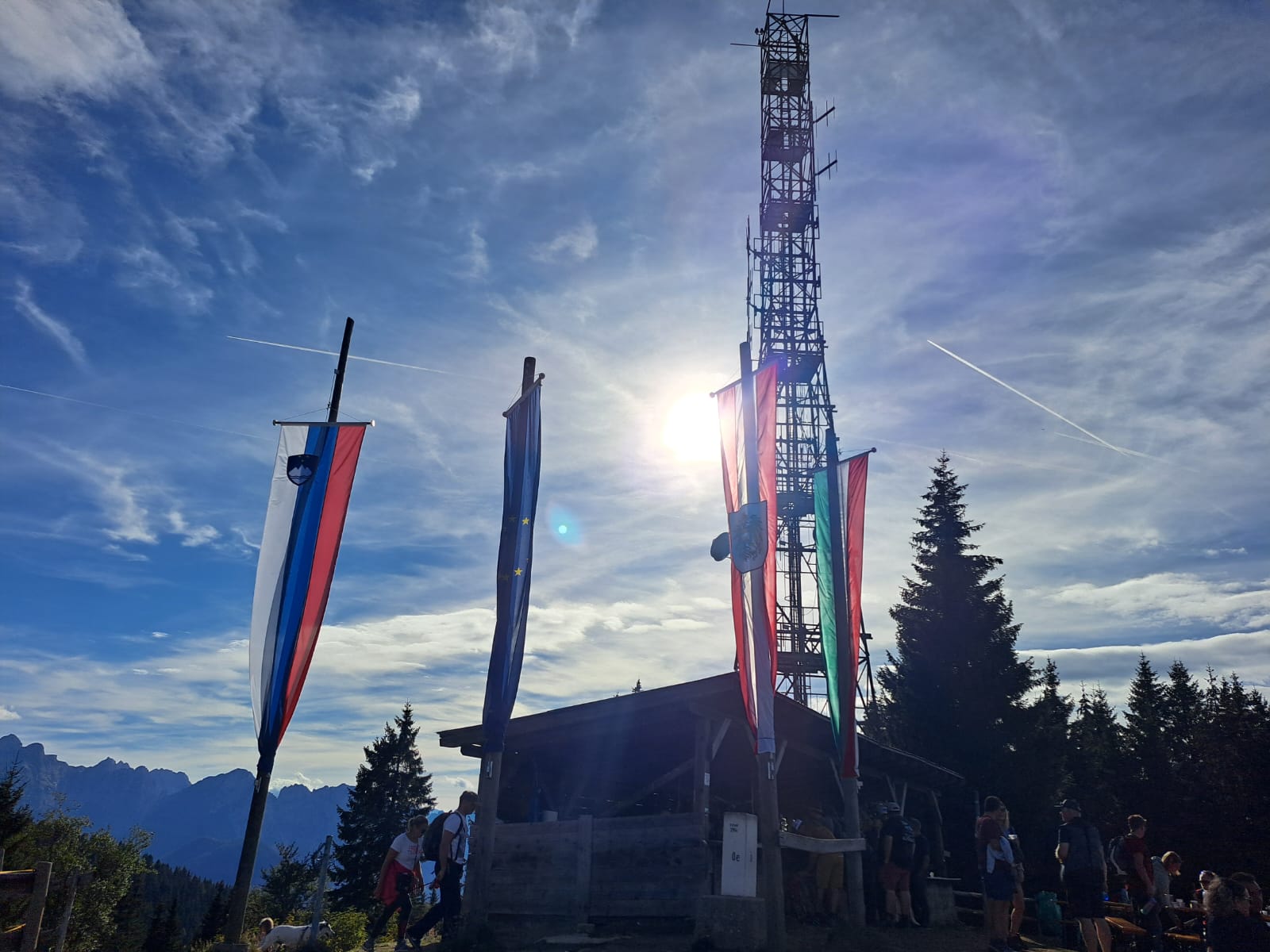 Bandiere di Slovenia, Unione Europea, Austria e Italia che sventolano sulla cima del Monte Forno sotto un cielo azzurro soleggiato.